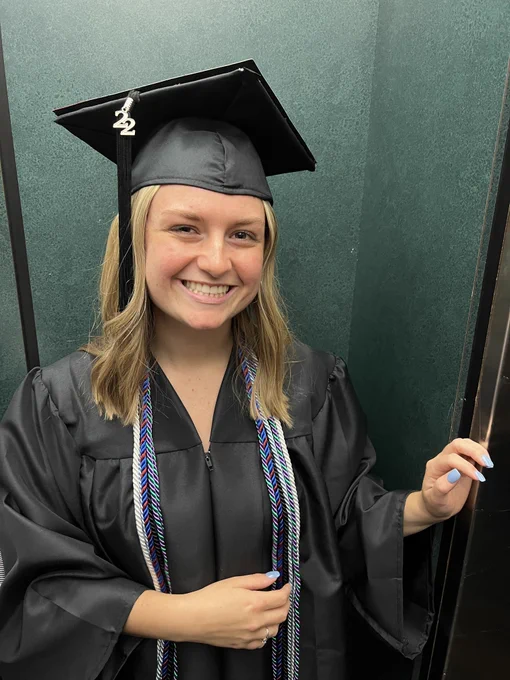 A smiling young woman in academic regalia standing in a doorway