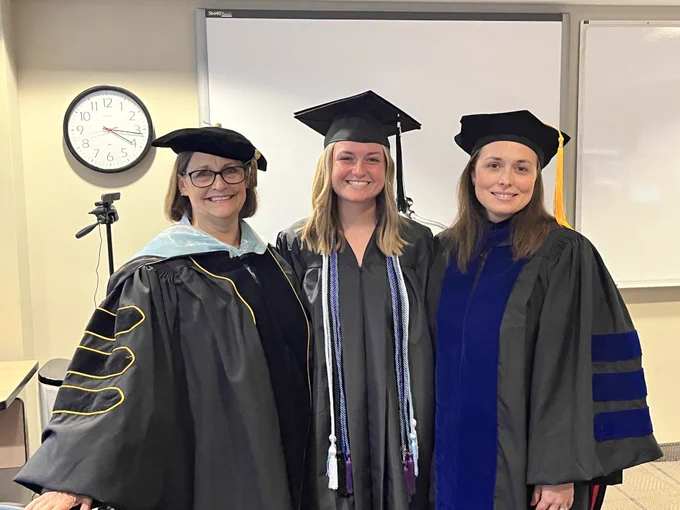 Two smiling women in doctoral regalia, with another in undergraduate regalia between them