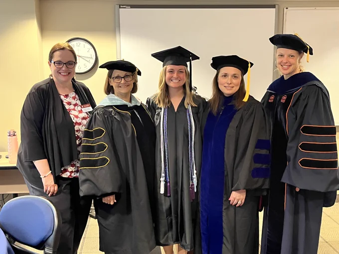 Five smiling women standing in a classroom, with four wearing academic regalia.