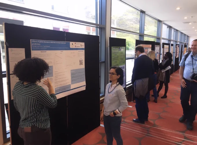 Photo of a corridor at the John McIntyre Conference Centre (Edinburgh) with academic posters pined on posterboards and lined up next to floor-to-ceiling windows. People are walking along the corridor, presenting posters, and talking to presenters.