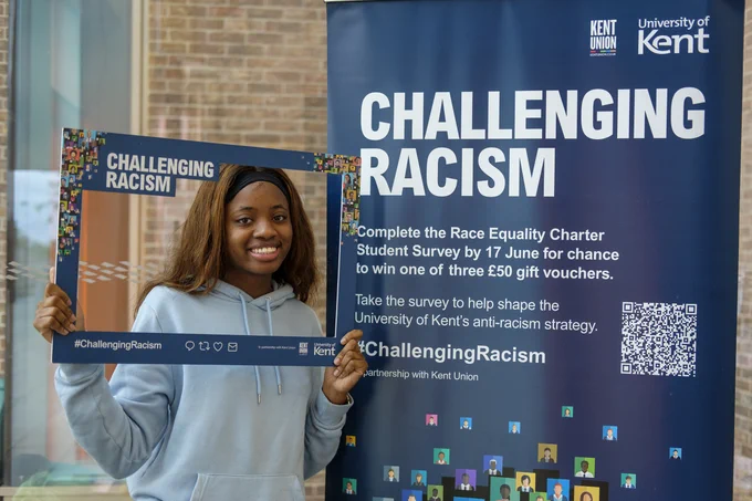 A Kent student posing and smiling whilst holding a challenging racism selfie frame. The student is standing next to a banner that reads 'Complete the Race Equality Charter Student survey by 17 June for a chance to win one of three £50 gift vouchers. Take the survey to help shape the University of Kent's anti-racism strategy."