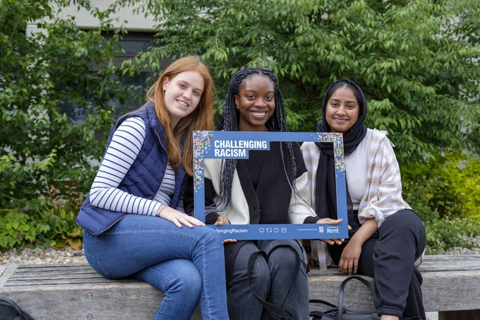 A group of Kent students smiling sitting on a bench holding the challenging racism selfie frame