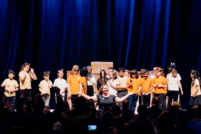 Children in animal costumes stand at the front of the stage ready to bow, with their Musical Director holding their arms wide facing the audience.