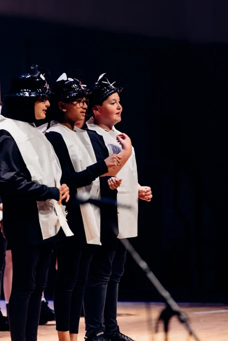 Three students sing on stage wearing white bibs and animals hats.