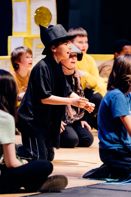 A young boy kneels on stage in front of a paper lighthouse wearing a top hat with ears attached, surrounded by his singing classmates.