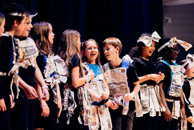 A group of children wearing costumes made of newspaper smile and sing on stage.