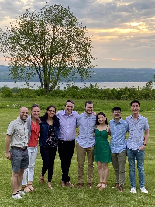 A group portrait shot of our friend group standing side-by-side in front of a scenic backdrop of Seneca Lake during the sunset. Eight of us are smiling together into the camera while surrounded by green foliage, bushes, and trees.