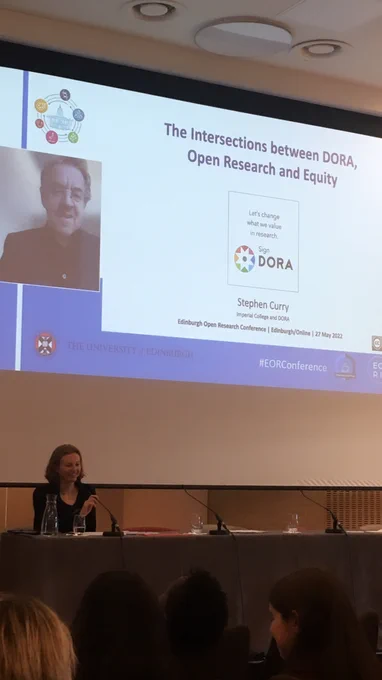 Photo showing the panel table and large screen inside the main conference room of the John McIntyre Conference Centre (Edinburgh). A young woman is sitting at the panel table. On the large screen behind her is, on the left-hand side, the camera of remote keynote speaker Stephen Curry, and on the right-hand side the introduction slide of his presentation.