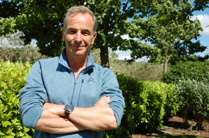 Robson Green pictured in a blue pullover jumper. In the background there are trees and hedges.