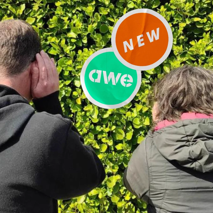 Image description: A man and a woman face away from the camera, towards a hedge with bright green leaves. They are cupping her ears, as if to listen closely. Graphics of stickers have been overlaid towards the top of the image, one showing the acronym ‘awe’ (for Art Week Exeter), the other showing the word ’new’. (Image credit: Gem Smith, Take-A-Part.)