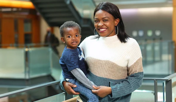 Rebecca Osamudiame, a Black woman with medium-length hair, stands next to a balcony in the Rowe building. She is wearing a white, blue and beige dress and gold hoop earrings. In one arm, she holds her son, a Black toddler wearing jeans and a blue shirt. She holds his hand with her free arm, and they both look at the camera, smiling.