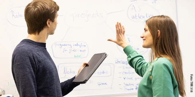 Mann mit Tablet-PC neben Frau, die in Richtung eines Whiteboards deutet