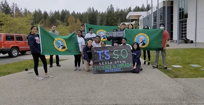 A group of students stand outside their school while holding a sign that reads "TSSO Tesla STEM Science Olympiad Nats 2022."