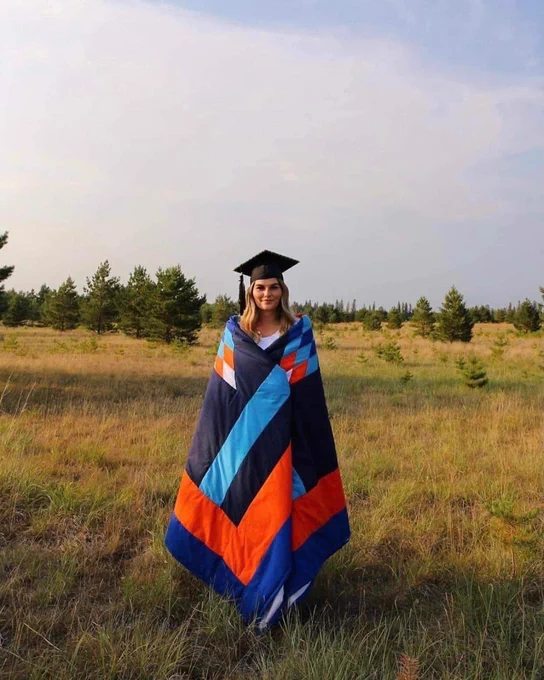 A Métis woman standing in a field with a star blanket and a grad cap on.