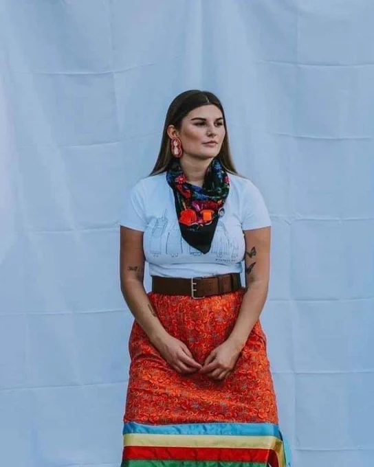 A Métis woman standing in a ribbon skirt against a white backdrop.