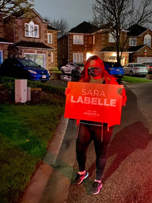 2022 Provincial ONDP Candidate for Whitby, Sara Labelle stands along a curb in Whitby holding an orange sign with their name written in white writing.There are houses with white windows and cars behind them. The picture is from May 4, 2022 when the writ dropped for the election.
