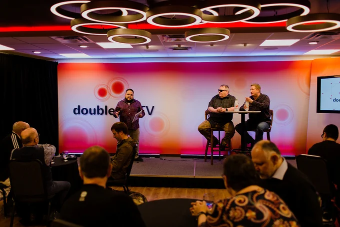 Marc stands on a stage with the Double Tap TV logo behind him in red, white and pink, with Steven and Shaun sat to his right. An audience is in the forefront, slightly blurred. 