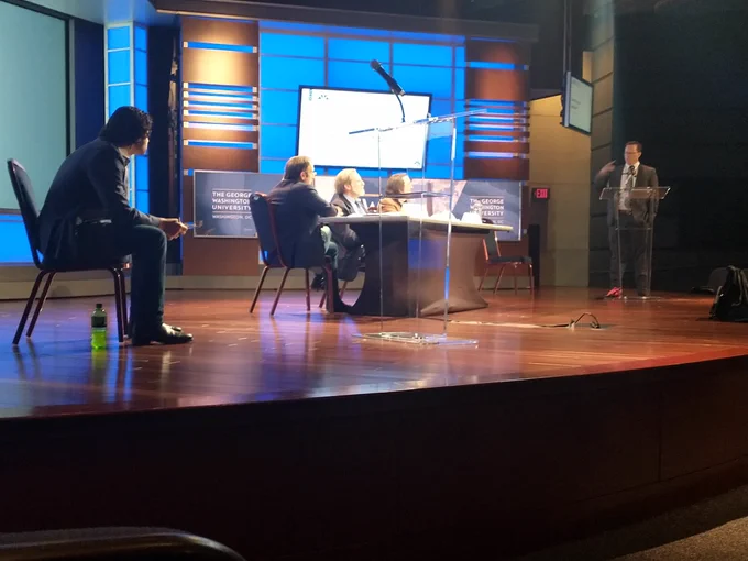 A stage featuring three men at a table, one man nearby sitting and taking notes, and another man at the far end of the stage arguing at a lectern. 