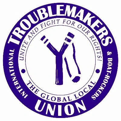 a circular blue and white badge with an illustration of a slingshot in the middle. the text on the outer blue ring reads "international TROUBLEMAKERS and boat-rockers UNION" - and in an inner ring of text, Unite and Fight For Our Rights - The Global Local