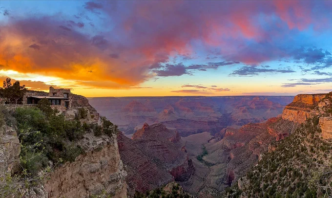 a rustic stone building on the edge of a cliff looking over a mile deep canyon landscape of buttes and cliffs. At sunset, brilliant yellow, orange and red clouds fill the sky. NPS photo/M. Quinn 06/10/2022