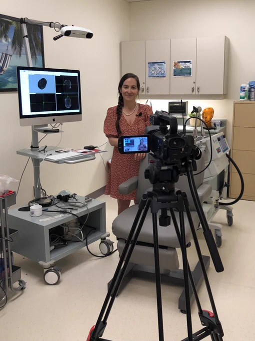 A video camera is focused on Dr. Sara Tremblay who stands next to a computer displaying images of the brain