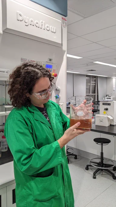 A man with curly black hair in a green labcoat holds up a large beaker containing about 1 L of brown liquid 
