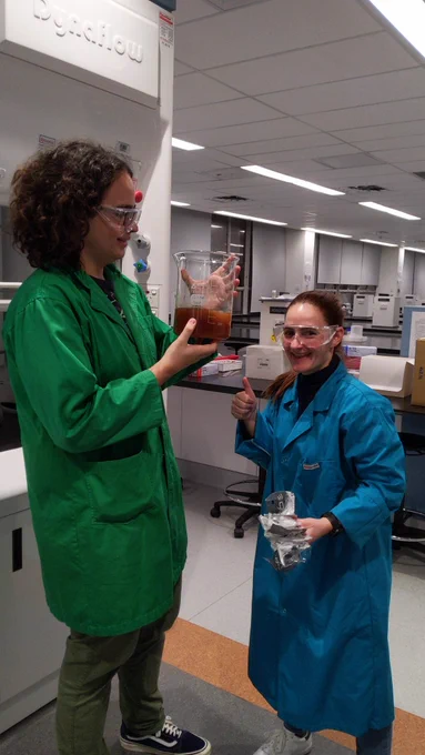 A man with curly dark hair in a green labcoat holds up a large beaker with about 1 L of brown liquid. Beside him, a woman in a turquoise labcoat gives the camera a thumbs up.
