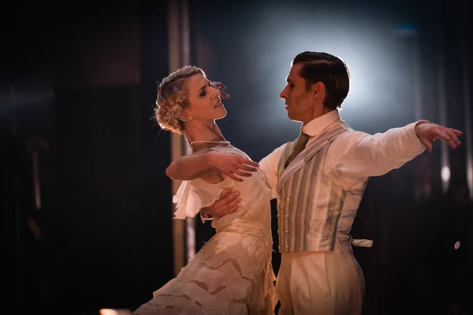 Antoinette Brooks-Daw and Ashley Dixon in The Great Gatsby. Both dancers are dressed in white and are in the centre of the image, highlighted by the stage lights in the background. Ashley has one arm around Antoinette's waist. Photo by Riku Ito.