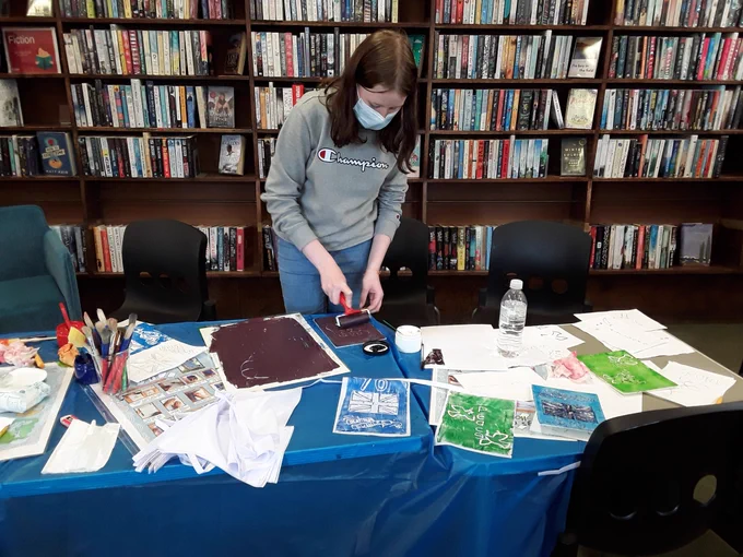 A young library volunteer rolls paint onto a piece of foam in preparation to print onto a flag. Table covered with paint pots, brushes and other flags 