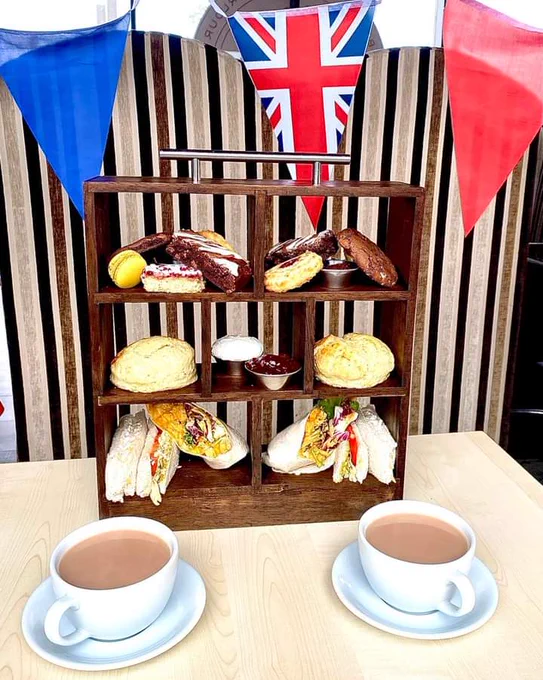 Afternoon tea stand with cakes and sandwiches, next to two cups of tea. Bunting in the background. 