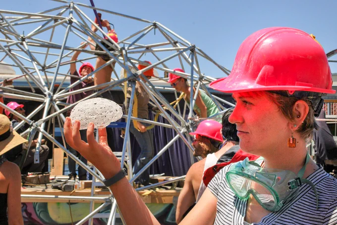 A woman wearing a pink hard hat and safety goggles looking at a small model of a brain while a large structure is built behind her