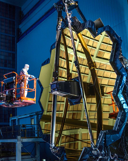 Webb's golden mirror is reflecting the light in a dim room with a blue background.  A person in a cleanroom suit stands on an orange lift in front of the mirror. 