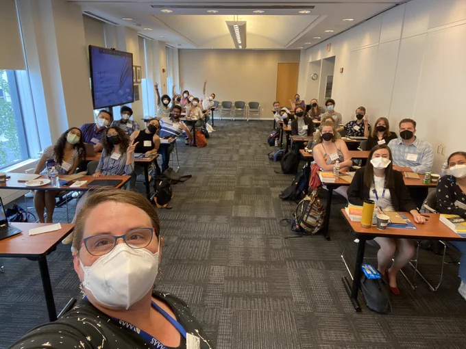 A masked selfie of the AAAS Mass Media Fellows and Diverse Voices Interns at their orientation in a classroom setting.. The fellows are in desks of two, and several fellows in the back are raising their hand.
