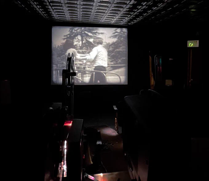 A black and white super8 film is being projected onto the screen showing a man and women on playground equipment. The projector can seen seen in the shot. 