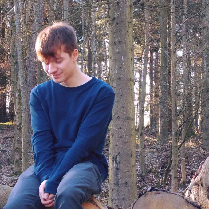 Sam is sat in the woods wearing a blue jumper and jeans. He smiles softly to himself.