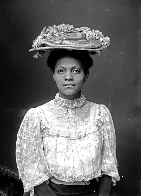 Unidentified African American woman in embroidered white shirt and straw hat with flowers sitting for a portrait. Her face is serious and dignified, her hands gathered in her lap. 