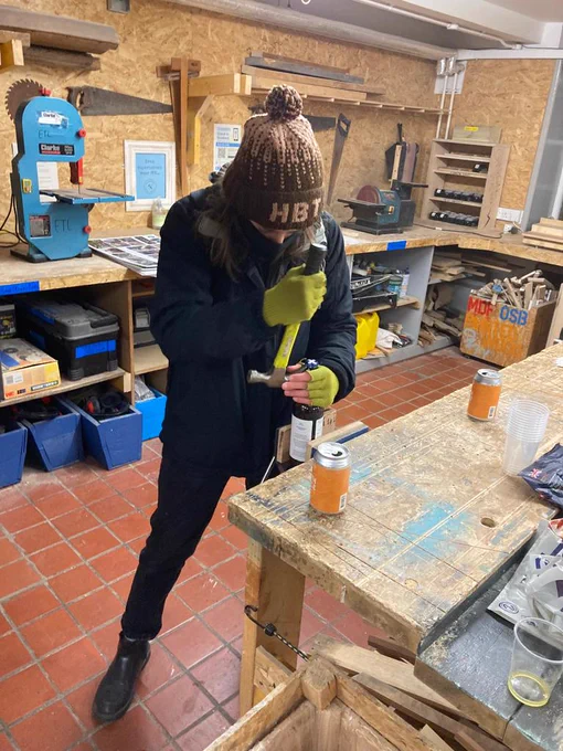 (ImageDescription: Jess, a volunteer with ETL, opens a can of beer with a claw hammer. She's wearing black clothes with a brown bobble hat and green work gloves)