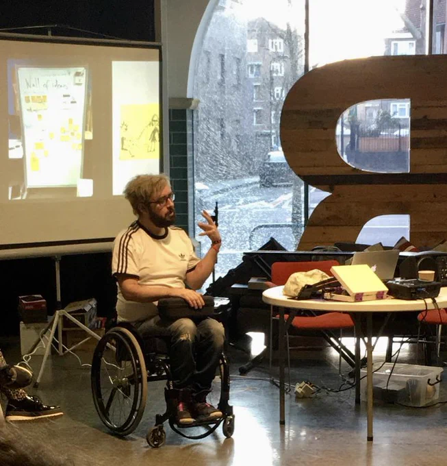 Associate Musician Kris Halpin discusses a project at a Drake Music Team Away Day. He sits in front of a screen on which images are projected and next to a table with equipment and tech.