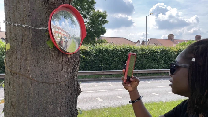 Woman standing next to a tree, you can see her face side on, only. She is holding up her phone and taking a photo of a tree trunk which has a round mirror attached to it. 