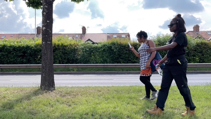 Two women walk along the grass verge next to a road. 
