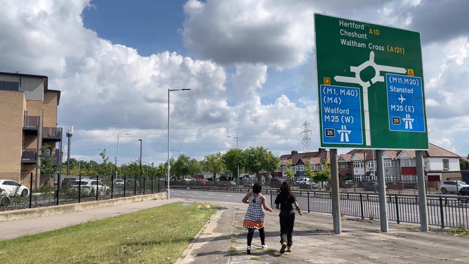 Two women walk towards a road sign for the M25 (West to Watford, East to Stansted). We see the back of them as they walk. A road on their right, some houses on the left. A small grass verge. Sunny