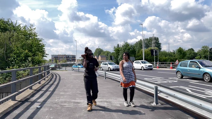 Two women walk towards us, along a busy main road. One woman has her phone to her ear, listening. 