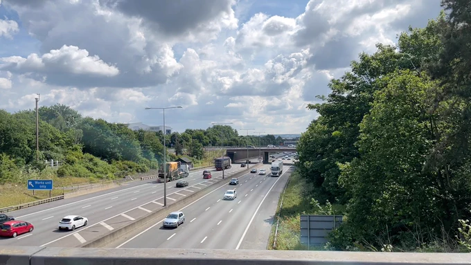 A view from a bridge over the M25. Three lanes of traffic each way. Cloudy white grey sky. Sunny