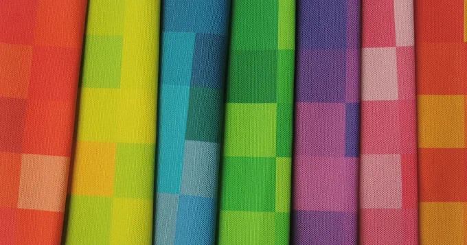 Selection of colourful fabric samples. From left to right the fabric samples  are coloured in a variety of shades of red, yellow, blue, green, purple, pink and orange. All of the fabric samples have a blocky design housing the solid gradient colour shifts.