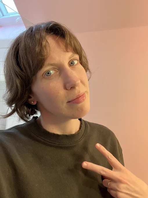 Selfie of a person in a green crew neck with shaggy short hair holding up 2 fingers in front of a pink wall