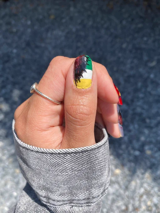 Stranger things season four nail art on a left hand in front of gray gravel. Close up of the green/white/gold stripes with the red cracks of the upside down portal on the left thumb.