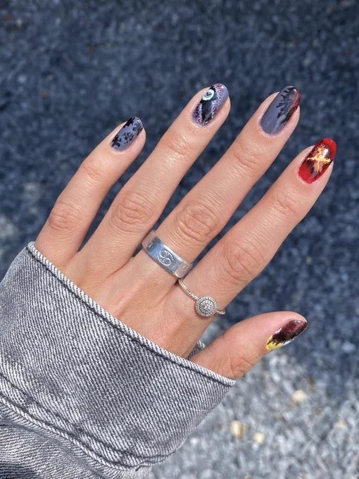 Stranger things season four nail art on a left hand in front of gray gravel. From pinky to thumb, gray background with black spiders, grandfather clock from the upside down on a gray background, a gray background of the upside down with a red crack that leads to Hawkins, the orange/yellow/red/black portal that leads to Hawkins, and green/white/gold stripes with the red cracks of the upside down portal.