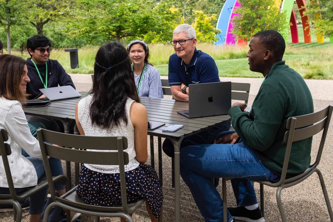 Tim, Susan Prescott, and five Swift Student Challenge winners sit at a table and talk outside at Apple Park.