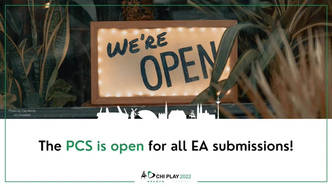 The picture shows a sign with the text 'we're open' framed in wood in a window full of green plants. The text below reads: The PCS is open for all EA submissions!