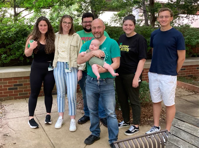 Six adults and one baby in front of some trees and a low brick wall. The adults look happy. The baby looks confused, probably because he doesn't know what's going on or why his father has taken him away from his toys.